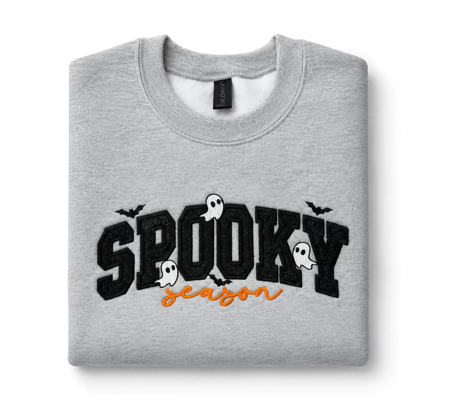 A folded light gray crewneck sweatshirt featuring a 'Spooky Season' graphic on the front with black block letters, white ghosts, and bats, and orange cursive text underneath, displayed on a clean white background.  Glitter Spooky applique, Gray Gildan crewneck sweatshirt with black glitter appliqué “Spooky Season” lettering, embroidered ghosts and bat details, folded flat lay