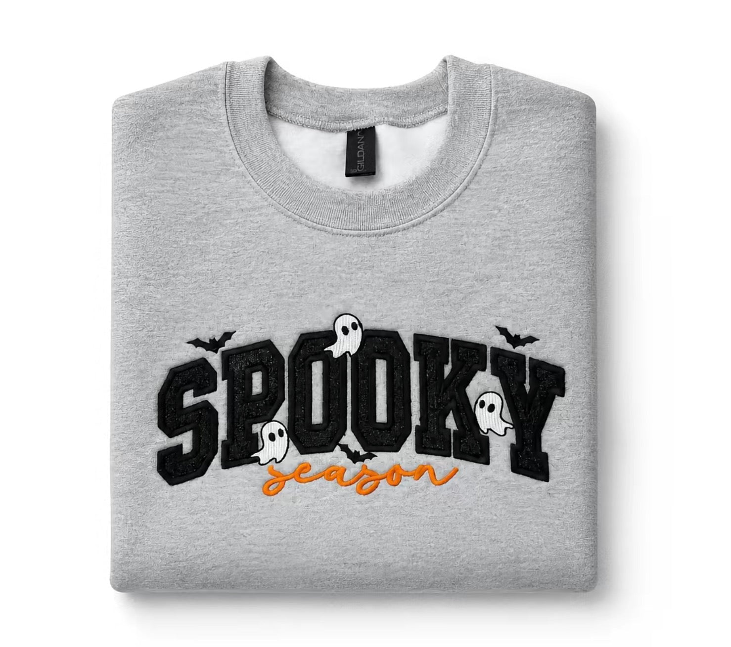 A folded light gray crewneck sweatshirt featuring a 'Spooky Season' graphic on the front with black block letters, white ghosts, and bats, and orange cursive text underneath, displayed on a clean white background.  Glitter Spooky applique, Gray Gildan crewneck sweatshirt with black glitter appliqué “Spooky Season” lettering, embroidered ghosts and bat details, folded flat lay