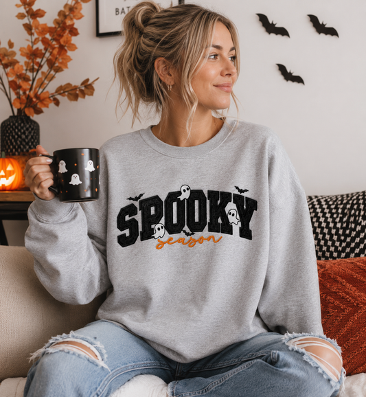 A woman sitting on a couch wearing a light gray crewneck sweatshirt with 'SPOOKY season' printed in black glitter appliqué and orange embroidery holding a black mug with ghost illustrations.