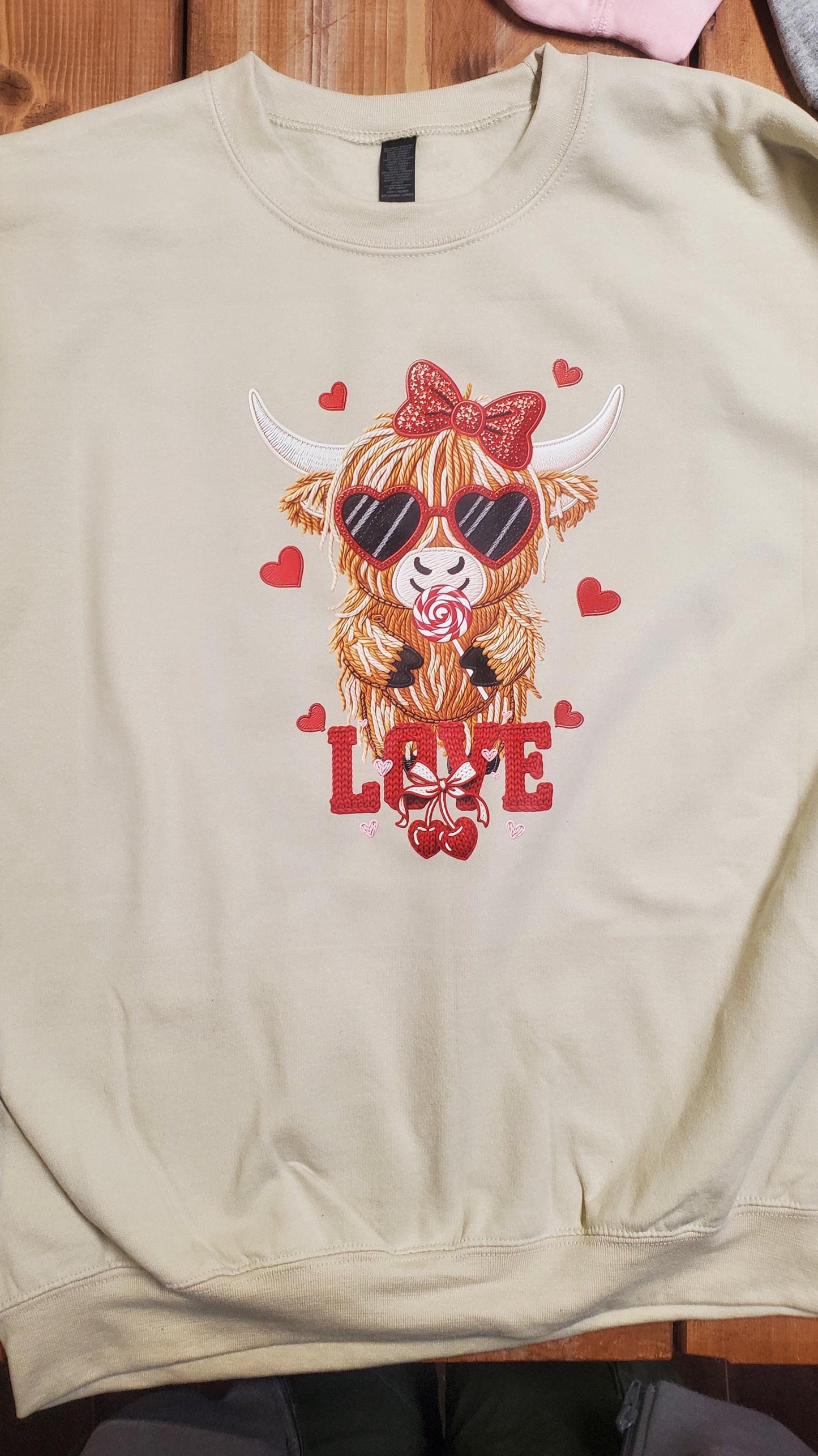 Cream colored sweatshirt featuring a cute Highland cow wearing heart sunglasses and a bow, holding a lollipop, surrounded by red hearts and the word love in a faux crochet yarn Valentine style, Angela Jasmina, AJ Blanks
