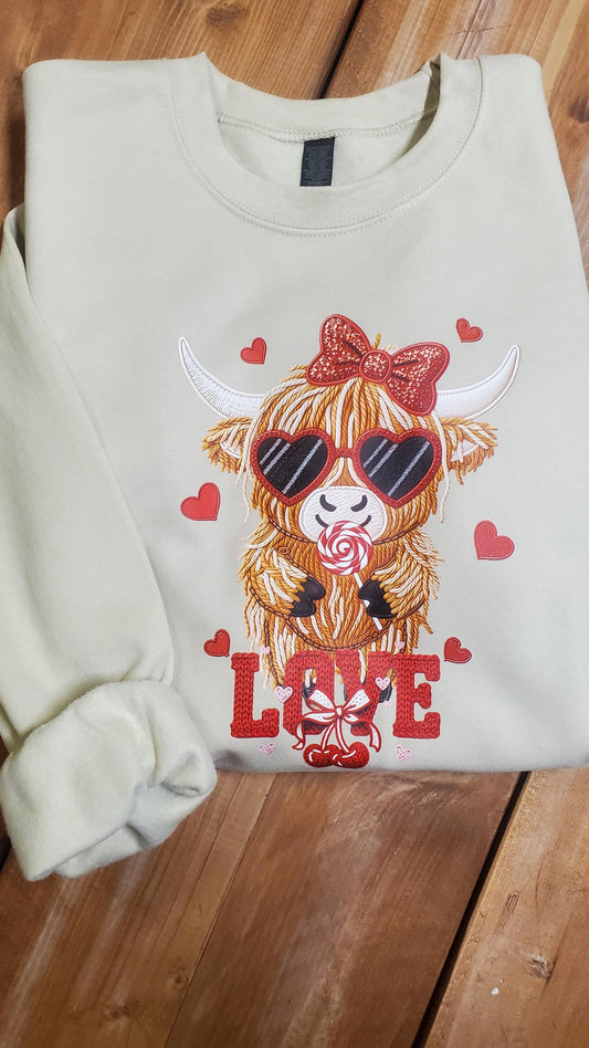 Cream colored sweatshirt featuring a cute Highland cow wearing heart sunglasses and a bow, holding a lollipop, surrounded by red hearts and the word love in a faux crochet yarn Valentine style, Angela Jasmina, AJ Blanks